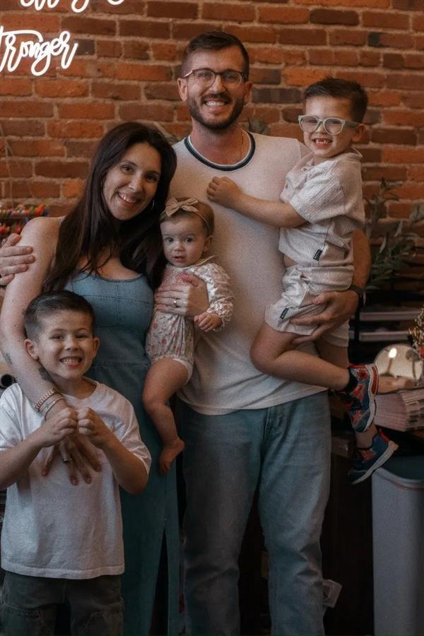 Five people posing indoors in front of a brick wall; one adult holds a baby