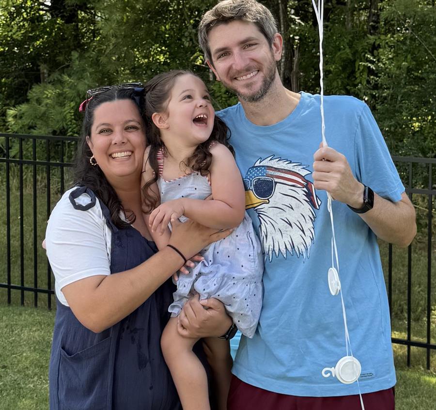 Two adults and a child smiling outdoors; one adult holds the child
