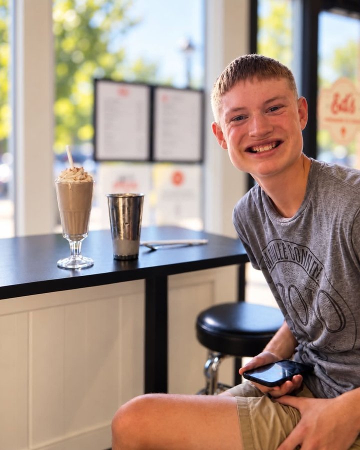 Young man smiling at a shop counter, holding a phone, beside a chocolate milkshake and metal mixing cup
