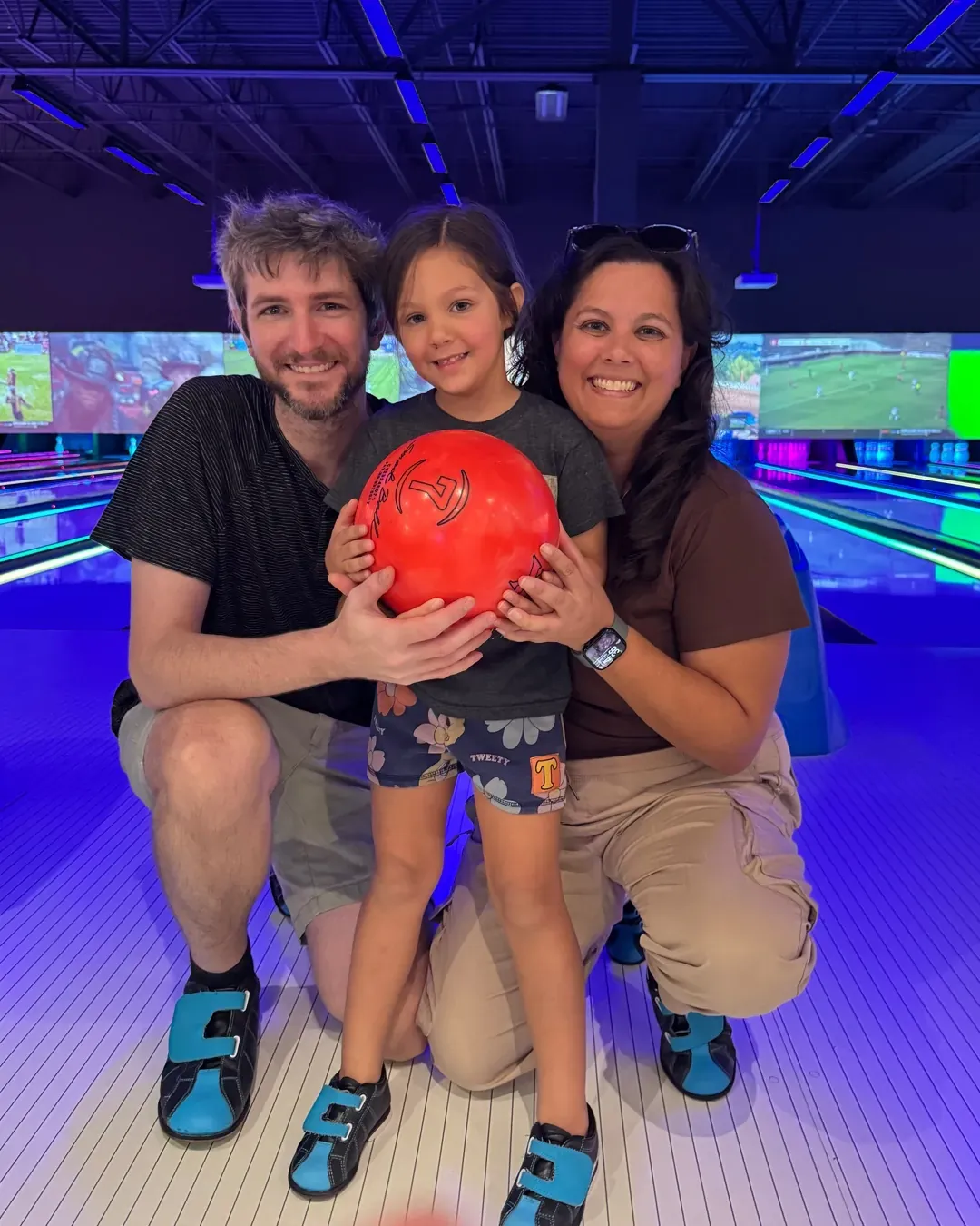 Dr. Taylor Hickok, her husband, and their daughter kneeling together at a bowling alley, holding a red bowling ball.
